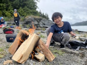 A young person feeds a flaming campfire on a sandy beach.